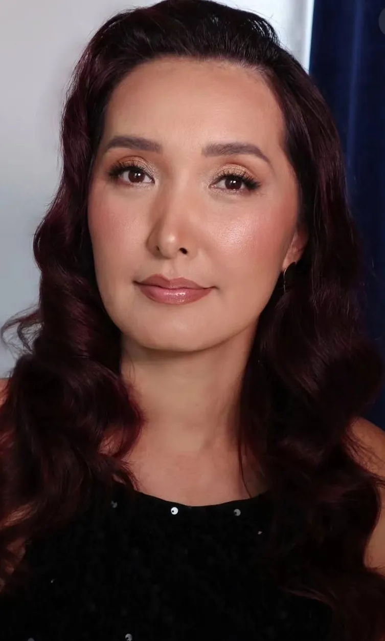 Portrait of a woman with deep burgundy waved hair, soft gold eye makeup and a nude-rose lip.