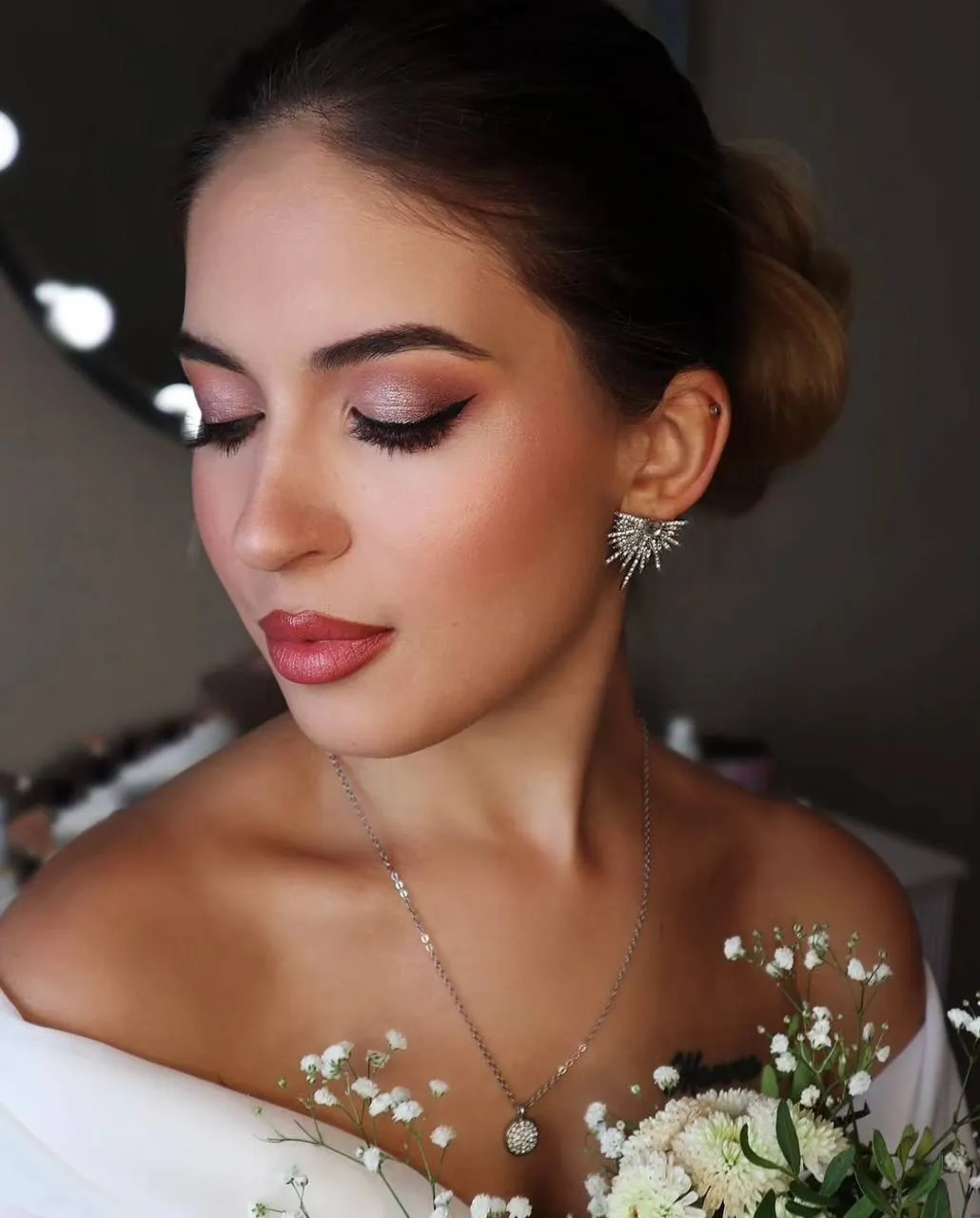 Bride with a low bun, soft rose eye makeup and a winged liner, holding white flowers.