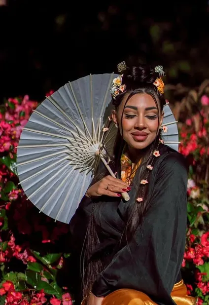 Portrait in a black and gold robe with a paper parasol, hair pinned with blossom, among pink flowers at night.
