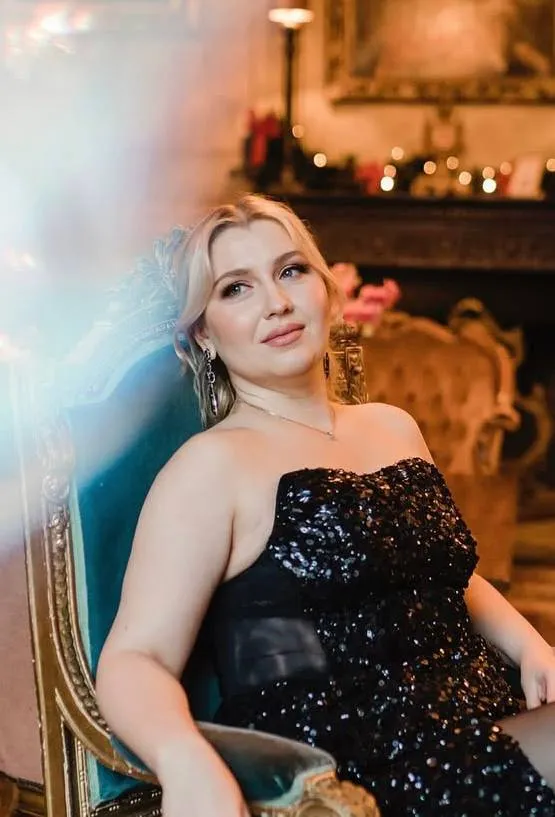 A blonde woman in a black sequin dress in a gilded armchair beside a lit fireplace, with soft neutral makeup.