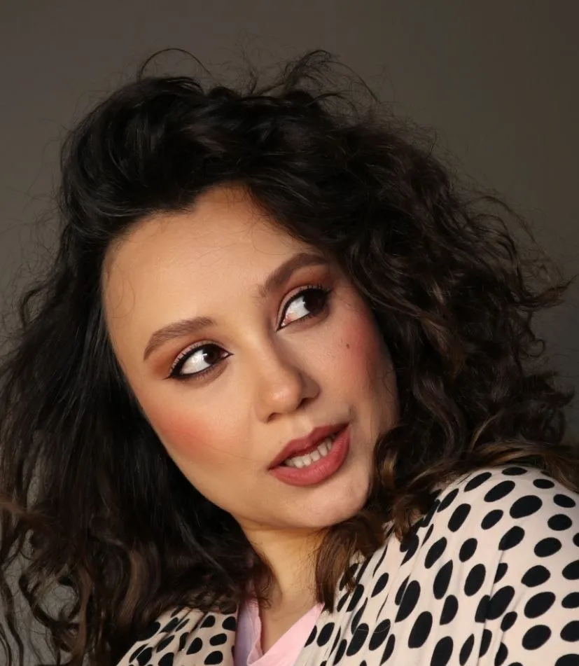 Studio portrait of a woman with full curly hair, soft peach eye makeup and a glossy lip.